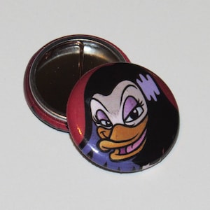 May include: A round button with a cartoon duck face design. The button has a red border and a shiny metal back. The duck has black hair, a purple and white face, and a yellow beak. The button is approximately 1.5 inches in diameter.