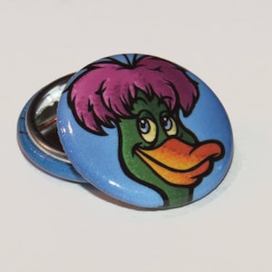 May include: A blue and white button with a cartoon duck wearing a pink and purple wig. The duck has a green head and a yellow beak.