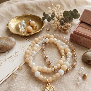 Op de afbeelding: Drie armbanden met kralen in perzik-, witte en gouden tinten, met een gouden hanger met een wit hart. De armbanden worden getoond met een gouden schaal met kralen, een klein bloemstuk en een kaart met "Handmade for You".