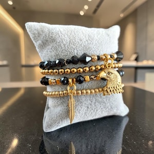 Op de afbeelding: Een set van vier gestapelde armbanden met zwarte gefacetteerde kralen, metallic goudkleurige bolvormige kralen en kleine goudkleurige kubussen. De armbanden bevatten een goudkleurige adelaarskopbedel en een goudkleurige veerbedel aan de strengen.