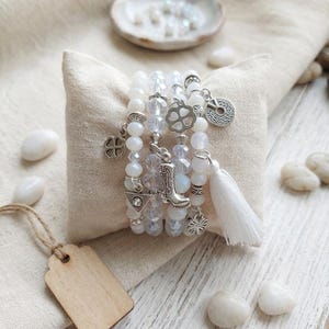 Op de afbeelding: Een stapel witte kralenarmbanden met zilveren bedels, waaronder een klavertjevier, een cowboylaars en een munt. Een witte kwast hangt aan een armband. De armbanden worden getoond op een klein, vierkant, beige kussen.