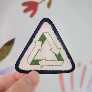 May include: A triangular sticker featuring a dark blue border and a white center. Inside the triangle is a green recycling symbol composed of three arrows arranged in a continuous loop, with a textured, stamped appearance on the green ink.