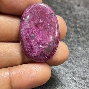Könnte beinhalten: Ein oval geschliffener Edelstein in tiefem Magenta- und Rosaton. Die Oberfläche weist eine natürliche, kristalline Textur mit sichtbaren inneren Einschlüssen, lichtreflektierenden Facetten und kleinen dunklen Mineralflecken im gesamten Stein auf.