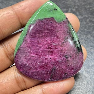 Könnte beinhalten: Ein polierter, birnenförmiger Rubin-Zoisit-Edelstein-Cabochon mit einem leuchtend magentafarbenen Zentrum, das von einem hellen lindgrünen Rand umgeben ist, mit kleinen schwarzen mineralischen Einschlüssen auf der glatten, reflektierenden Oberfläche.