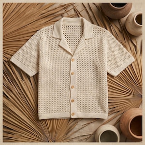 May include: Cream-colored crochet shirt with short sleeves and a collared neckline. The button-down shirt features wooden buttons and a breathable, open-weave design. The shirt is displayed on a neutral background with dried palm leaves and pottery.