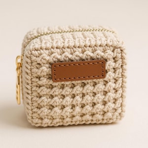 May include: A small, square, beige crochet pouch with a textured, hand-knitted appearance. It features a gold-toned metal zipper closure on the top and a rectangular, smooth brown leather-like patch centered on the front face of the item.