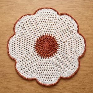 May include: A handmade, crocheted doily in a flower shape. The doily features a white open-weave center with a rust-colored center circle and a rust-colored scalloped edge. The doily is placed on a light brown wooden surface.