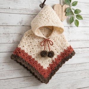 Peut inclure: Un poncho à capuche de couleur crème avec un motif en crochet texturé. Le vêtement comprend un cordon de serrage avec deux pompons marron foncé aux extrémités. Le bord inférieur est fini par des rayures horizontales orange rouille et marron.