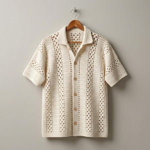 May include: Cream-colored crochet shirt with short sleeves and a collared neckline. The shirt features a button-down front and a textured, open-weave design. The shirt is hanging on a wooden hanger against a neutral background.