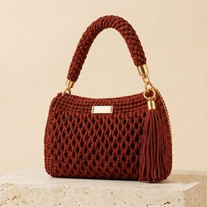 May include: A dark rust-colored crochet handbag featuring a textured open-weave pattern, a thick braided handle, and a gold-toned metal plaque on the front. The bag is accented with a matching long tassel and a gold-toned chain strap attachment.