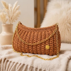 May include: A brown, crocheted handbag with a gold chain strap and a gold circular clasp. The bag is rectangular with a curved top and is resting on a cream-colored blanket. A vase with dried flowers is in the background.