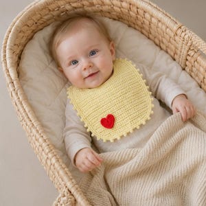 May include: A pale yellow crocheted baby bib featuring a small, vibrant red heart appliqué centered near the bottom. The bib has a textured, ribbed stitch pattern and a decorative picot edge trim, providing a soft and detailed appearance.