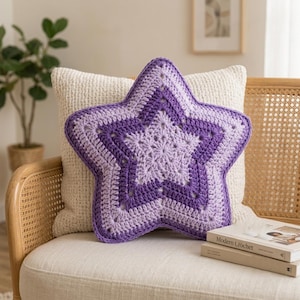 Può includere: Un cuscino all'uncinetto a forma di stella caratterizzato da un design a strati in tonalità lavanda chiaro e viola intenso. Il cuscino ha un aspetto testurizzato e cucito a mano con un motivo a stella centrale e un bordo spesso in filato viola.