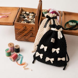 May include: A wooden sewing box with a handle, filled with buttons, sits next to spools of thread and decorative ribbons. Two drawstring bags, one black with cream bows and one cream with a black bow, are also present.