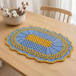 May include: An oval-shaped crochet doily featuring a central rectangular pattern in bright yellow, surrounded by concentric rings of light blue and yellow yarn. The edges of the doily are finished with a scalloped border in yellow yarn.