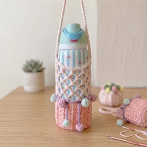 Puede incluir: Una botella de agua a rayas azul claro y blancas con tapa azul claro, dentro de un soporte de ganchillo hecho a mano. El soporte tiene una red color crema, una base rosa claro y pequeños pompones decorativos en azul claro y lavanda.