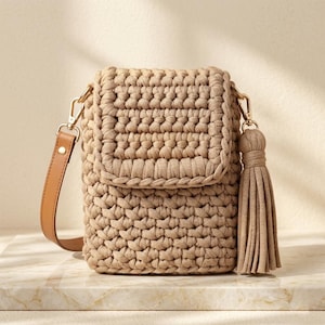 May include: A beige crochet crossbody bag with a flap closure and a brown leather strap. The bag features a decorative tassel and gold-colored hardware. The bag is made of a thick, textured yarn.