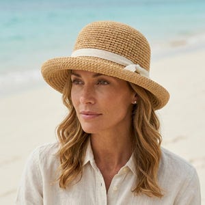 May include: A light brown woven straw hat with a rounded crown and a wide brim. It has a cream-coloured ribbon band and a small bow. The hat is made of natural fibres. The background shows a beach scene with the sea in the distance.