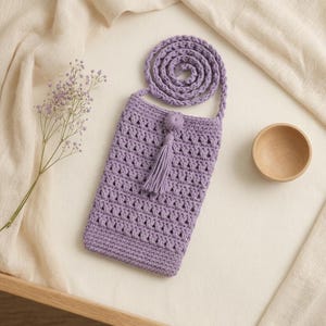 Può includere: Una borsa a tracolla all'uncinetto color lavanda con corpo rettangolare, motivo a punto traforato, base solida e una lunga tracolla abbinata. La borsa è decorata con una nappa centrale fissata a un piccolo bottone rotondo all'uncinetto.