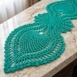 May include: A teal crochet table runner with a detailed openwork design. The runner is made of soft yarn and features a repeating pattern of circular and leaf-like shapes. The runner is placed on a marble-patterned surface.