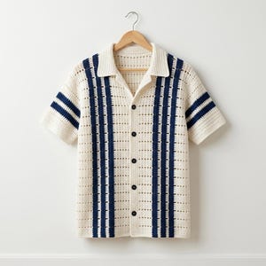 May include: Cream-colored, short-sleeved crochet shirt with navy blue stripes. The shirt features a collared neckline and a button-down front. The sleeves have navy blue stripes. The shirt is hanging on a wooden hanger.