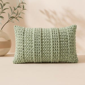 Pode incluir: Uma almofada retangular de malha verde sálvia com uma superfície texturizada. A almofada apresenta uma combinação de padrões verticais canelados e tecidos. A almofada é um elemento decorativo para casa.