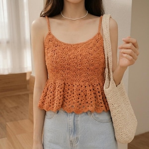 Op de afbeelding: Een oranje gehaakte top met spaghettibandjes en een uitlopende peplumzoom. De top wordt gecombineerd met lichtblauwe jeans. Een crèmekleurige gehaakte totebag is zichtbaar.