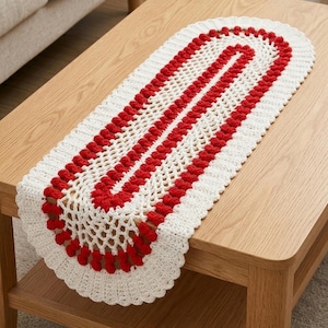 May include: Oval-shaped, hand-crocheted table runner. The runner features a white base with a red, textured stripe design down the center. The edges are scalloped and trimmed with red bobbles. The runner is displayed on a light-colored wooden table.