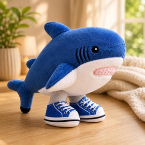 May include: A plush shark toy with a deep blue upper body and a white underbelly. The shark features black embroidered eyes and gill slits, a light pink mouth with visible teeth, and is wearing a pair of blue and white lace-up canvas sneakers.