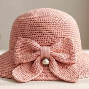 May include: A light pink, crocheted sun hat with a wide brim and a large bow detail. The bow is centered on the front and features a pearl-like embellishment. The hat has a rounded crown and a textured appearance.