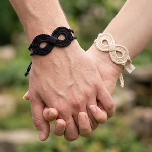 May include: A black cord bracelet and a cream-colored cord bracelet, each featuring a central infinity symbol design made of coiled rope. The cream bracelet includes a small rectangular tag attached to the side of the infinity knot.