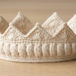 Puede incluir: Una corona de ganchillo de color crema con un diseño texturizado. La corona presenta picos puntiagudos y una fila de puntadas decorativas en forma ovalada a lo largo de la base. La corona hecha a mano es probablemente para un niño o para fines decorativos.