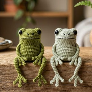 May include: Two handmade crochet frog figurines. One is olive green, the other is light gray. Both have large black eyes and are sitting with their legs extended. The frogs are made of yarn and are on a wooden surface.