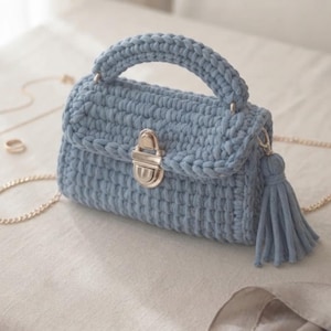 May include: A light blue crochet handbag featuring a thick top handle, a silver-toned metal push-lock closure on the front flap, and a matching blue yarn tassel attached to the side. The bag includes a thin gold-toned metal chain shoulder strap.