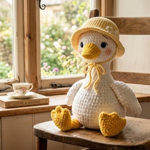 Puede incluir: Una muñeca de ganso de ganchillo hecha a mano con un sombrero y pies amarillos, y un lazo amarillo. El ganso es blanco con un pico amarillo y mejillas rosadas. Está sentado en una silla de madera cerca de una ventana.