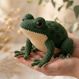 Op de afbeelding: Een handgemaakte gehaakte kikker in groen en beige. De kikker heeft grote, zwarte ogen en gedetailleerde kenmerken. Het amfibie is een zachte sculptuur, perfect als speelgoed of decoratie.