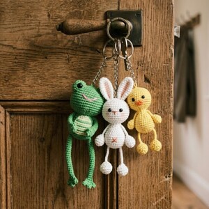 May include: Three crocheted keychains hang from a metal ring. A green frog, a white bunny with pink-lined ears, and a yellow duckling are the subjects. The keychains are attached to a wooden door with a metal handle.