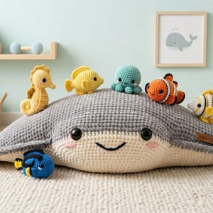 May include: A large, gray and cream-colored crochet stingray plush toy with a smiling face, featuring several smaller crocheted sea creature toys resting on top, including a yellow seahorse, a yellow fish, a teal octopus, and orange and white fish.