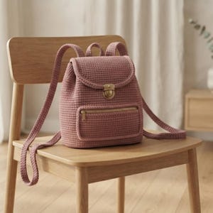 May include: A dusty rose crochet backpack featuring a structured flap with a brass-toned metal push-lock closure, a front zippered pocket with a matching brass-toned zipper, and two adjustable shoulder straps made from the same textured yarn.