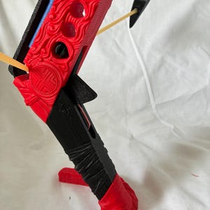 May include: A red and black crossbow with detailed carvings. The bow has a decorative design on the red body, a black handle, and a yellow arrow. The crossbow is set against a white background.