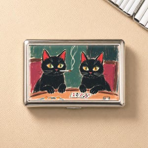 Könnte beinhalten: Eine silberne Zigarettenetui mit einer Illustration von zwei schwarzen Katzen, die Poker spielen. Eine Katze raucht eine Zigarette, die andere hat Karten. Der Hintergrund ist grün mit roten Akzenten.