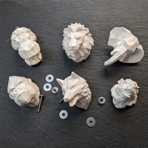Könnte beinhalten: Ein Set aus sechs weißen, geometrischen Möbelknöpfen in Tierform, darunter ein Löwe, ein Elefant und weitere Wildtiere. Das Set enthält metallische Befestigungselemente wie Schrauben und Unterlegscheiben für die Montage.