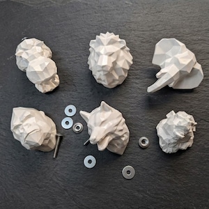 Può includere: Una collezione di pomelli per mobili a forma di animali geometrici in grigio chiaro. I pomelli includono un leone, un lupo, un elefante e altre forme astratte. Sono presenti anche viti e rondelle.