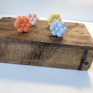 Peut inclure: Quatre boutons de placard en forme de fleur, orange, blanc, jaune et bleu, posés sur une boîte en bois rustique. La boîte présente des renforts d'angle en métal et une finition vieillie. Les boutons ajoutent une touche de couleur.