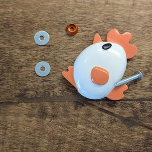 Peut inclure: Un bouton de meuble en forme de poulet blanc et orange avec un détail d'œil noir, accompagné de deux rondelles en métal argenté, d'une petite entretoise conique orange et d'un boulon de fixation argenté pour tiroir ou porte.