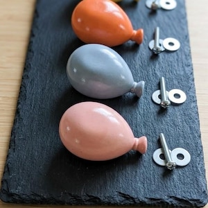 Peut inclure: Quatre objets colorés en forme de ballon, jaunes, oranges, gris et roses, sont disposés sur une ardoise noire rectangulaire. Chaque ballon a une petite tige et est associé à une vis et une rondelle. L'ensemble est posé sur un fond en bois clair.
