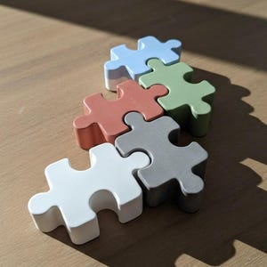 Peut inclure: Cinq pièces de puzzle épaisses et imbriquées dans des tons mats pastel et neutres. L'ensemble comprend une pièce bleu clair, une vert sauge, une terre cuite, une grise et une blanche, disposées en groupe sur une surface plane avec des ombres.