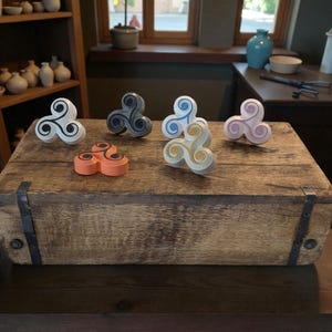 Op de afbeelding: Een verzameling van zes decoratieve, drielobbige ornamenten in verschillende kleuren, waaronder wit met zwarte accenten, grijs, oranje met zwarte accenten, blauw en geel, en roze en geel. Ze worden tentoongesteld op een rustieke houten kist.
