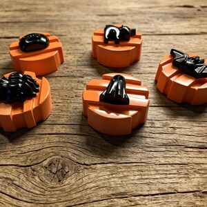 Puede incluir: Cinco bases circulares de color naranja con forma de calabaza, cada una rematada con una figura negra distinta. Las figuras incluyen una araña, un fantasma, un murciélago, un gato y una forma ovalada lisa. Cada base naranja presenta estrías verticales.