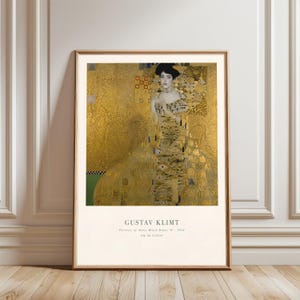 May include: A framed print of Gustav Klimt's "Portrait of Adele Bloch-Bauer I" from 1912. The artwork features a woman in an ornate gold dress against a gold background. The print is in a wooden frame and is displayed against a white wall.